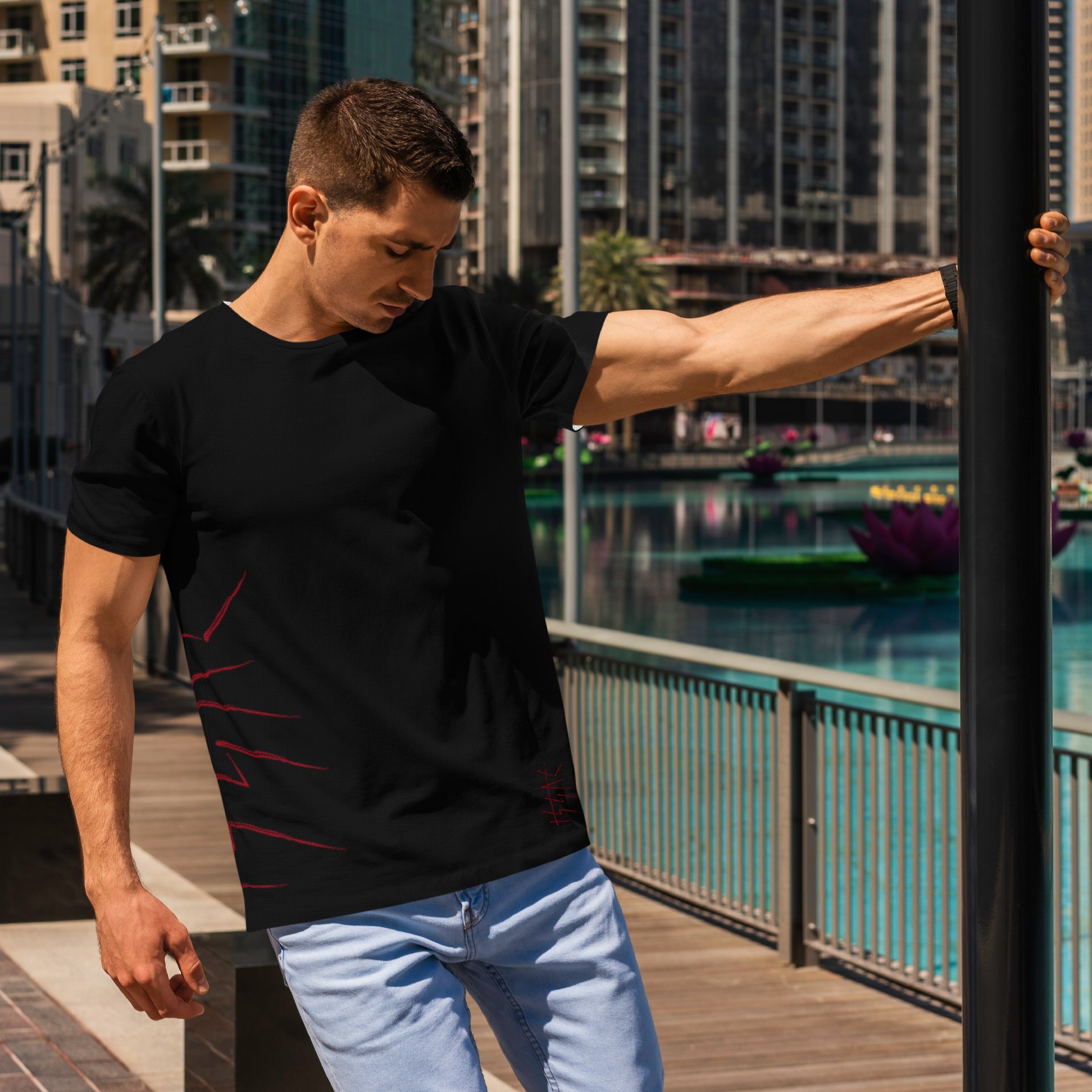 Man wearing a black t-shirt and light blue jeans standing on a waterfront promenade with buildings in the background.