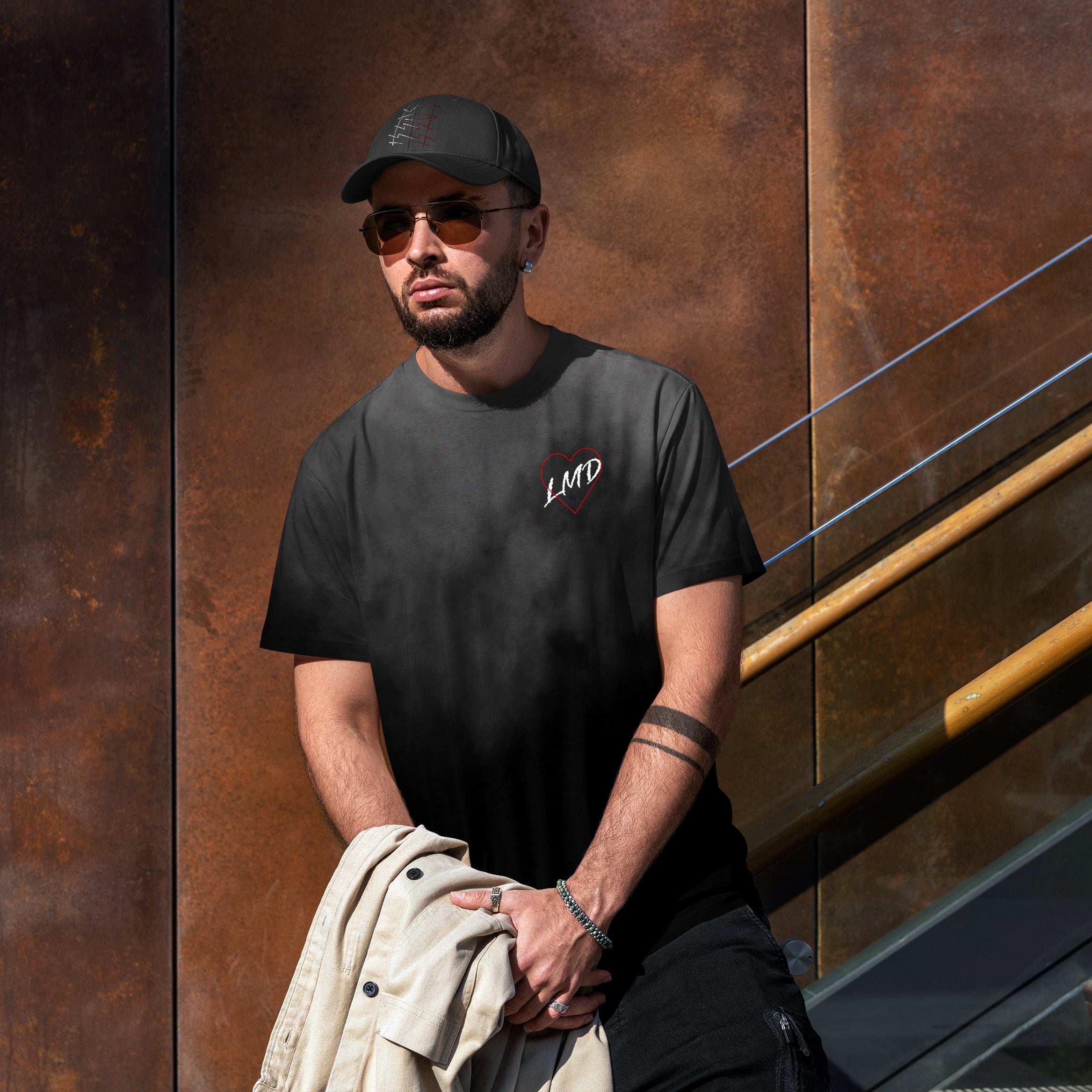 Man wearing a black t-shirt with 'LMD' logo, sunglasses, and cap standing against a metallic wall.