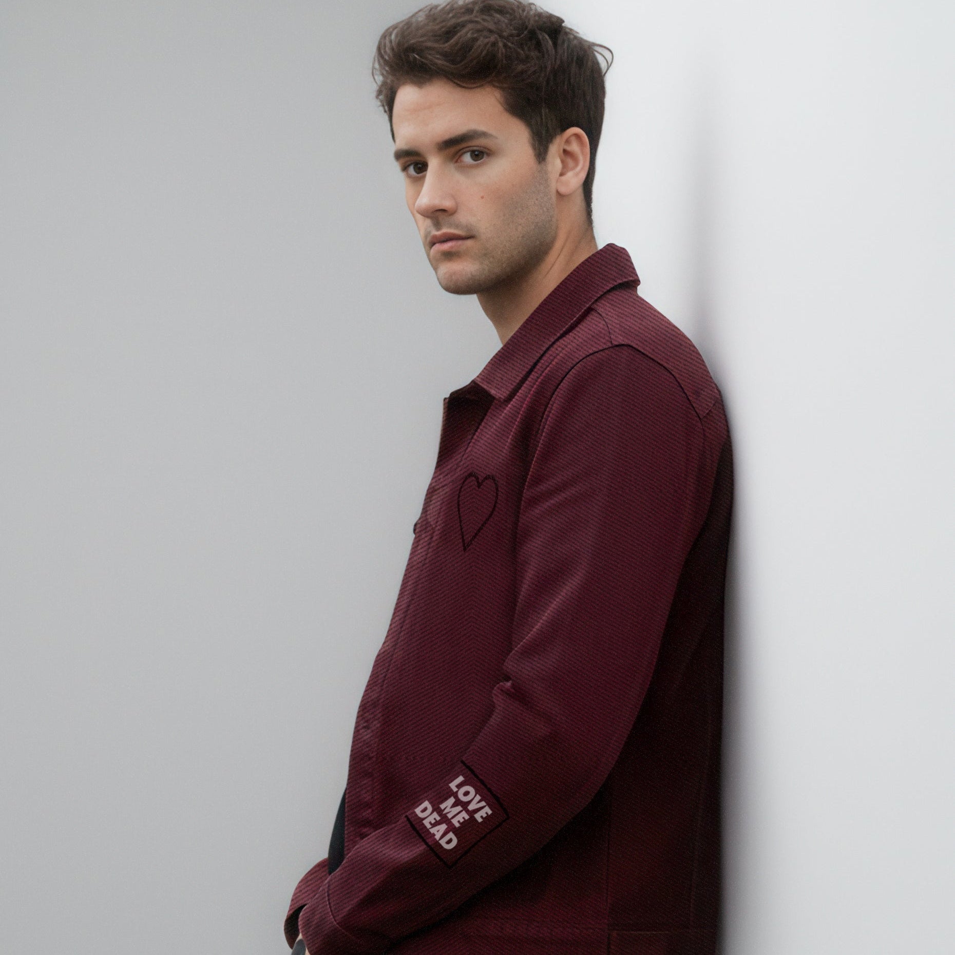 Man wearing a burgundy jacket leaning against a white wall.